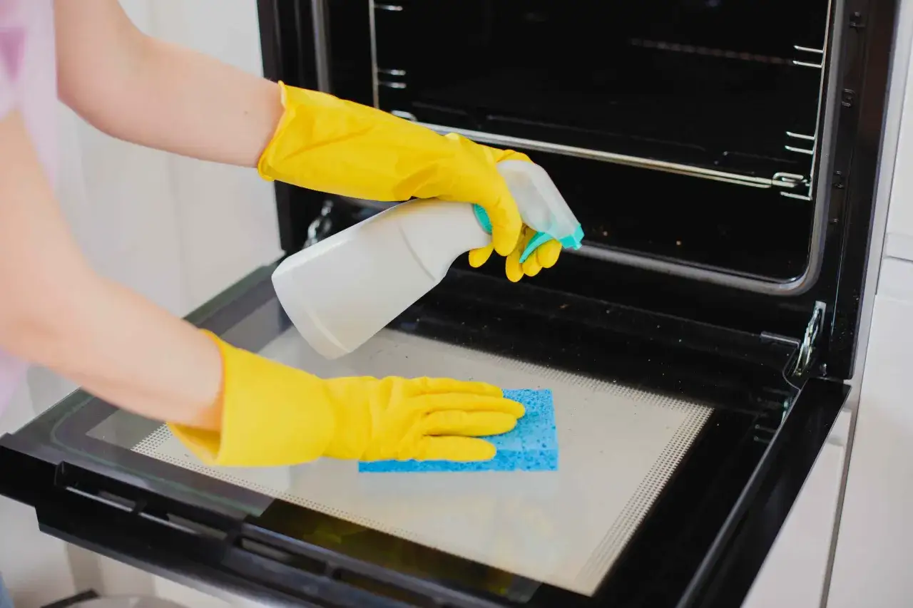 Manos con guantes amarillos rociando limpiador y frotando el cristal del horno con una esponja azul. Guía para como limpiar el horno.